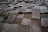 A textured surface composed of various square wooden tiles arranged in a grid pattern. Each tile has distinct grain patterns and shades, creating a three-dimensional appearance.