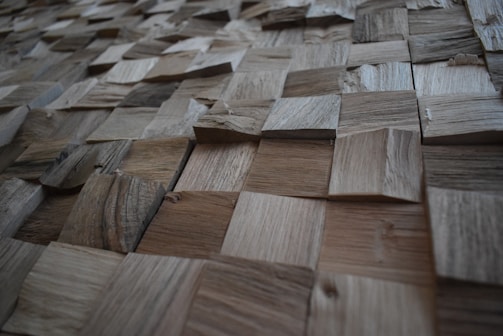 A textured surface composed of various square wooden tiles arranged in a grid pattern. Each tile has distinct grain patterns and shades, creating a three-dimensional appearance.