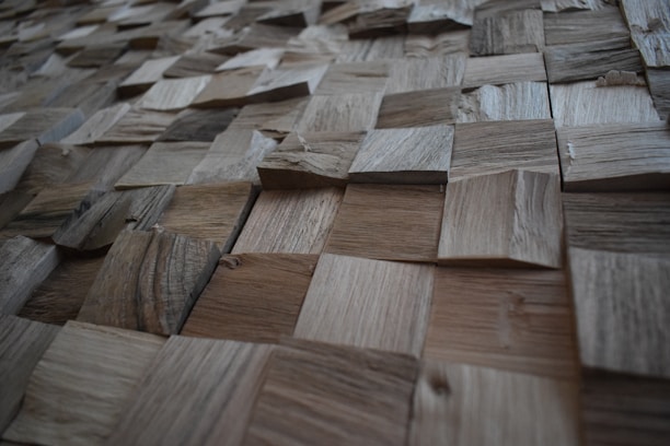 A textured surface composed of various square wooden tiles arranged in a grid pattern. Each tile has distinct grain patterns and shades, creating a three-dimensional appearance.