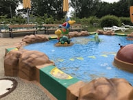 A small children's pool with colorful animal figures emerging from the water. The pool is surrounded by artificial rocks and green trim. Benches and lounge chairs are seen in the background, with trees and shrubs adding a natural ambiance.