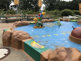 A small children's pool with colorful animal figures emerging from the water. The pool is surrounded by artificial rocks and green trim. Benches and lounge chairs are seen in the background, with trees and shrubs adding a natural ambiance.