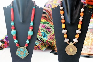 Two ornate necklaces are displayed on black stands. The left necklace features a large, blue and red geometric pendant with beads in turquoise and red. The right necklace displays a circular, patterned gold pendant with beads in white, orange, and brown hues. Both necklaces are set against a colorful, patterned fabric backdrop.