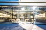 An Apple retail store with large glass windows and a prominent Apple logo above the entrance. Inside, there are wooden tables with electronic devices displayed along with colorful artwork on the walls. The bright, modern space is filled with natural light.