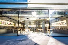 An Apple retail store with large glass windows and a prominent Apple logo above the entrance. Inside, there are wooden tables with electronic devices displayed along with colorful artwork on the walls. The bright, modern space is filled with natural light.