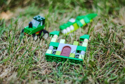 A small, colorful Lego tractor and a partially constructed Lego structure are placed on grass. The tractor is primarily green with black wheels and sits to the left. The structure consists of green and white bricks with a brown door, lying in the foreground.