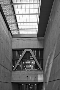 An architectural interior featuring concrete walls and a large window structure with triangular frames. The ceiling is composed of a grid of glass panels allowing natural light to illuminate the space. The overall design is modern and industrial, highlighting clean lines and geometric shapes.