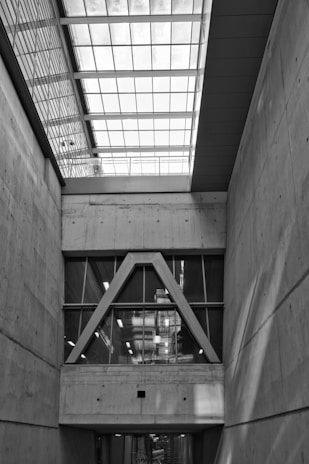 An architectural interior featuring concrete walls and a large window structure with triangular frames. The ceiling is composed of a grid of glass panels allowing natural light to illuminate the space. The overall design is modern and industrial, highlighting clean lines and geometric shapes.