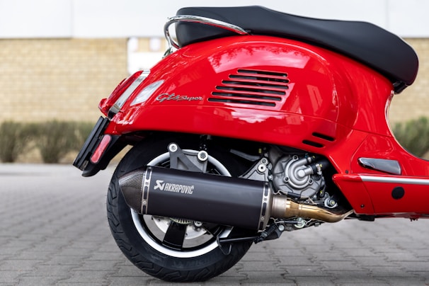 A close-up view of the back part of a red scooter, showcasing the detailed mechanics of the rear wheel, exhaust system, and engine components. The scooter has a shiny red paint finish and features the branding 'Akrapovic' on the exhaust.