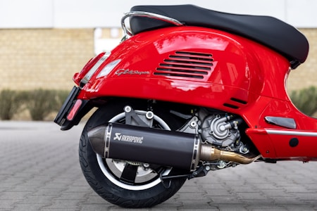 A close-up view of the back part of a red scooter, showcasing the detailed mechanics of the rear wheel, exhaust system, and engine components. The scooter has a shiny red paint finish and features the branding 'Akrapovic' on the exhaust.