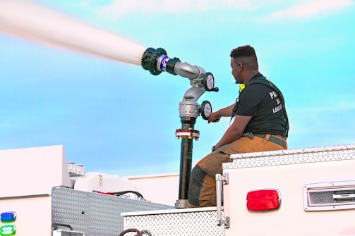 A person is seated on a fire truck, operating a fire hose nozzle that is actively spraying water. The person is wearing a dark shirt and brown overalls. The sky is clear and bright, suggesting daylight.