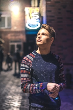 A young person is standing in a dimly lit urban alleyway, looking upward with a contemplative expression. The individual is wearing a dark sweater with colorful, intricate patterns on the sleeves. A neon sign is glowing brightly in the background, casting light on wet cobblestones and surrounding brick buildings.