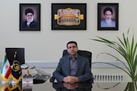 A man sits at an office desk with a reflective surface. There are three framed pictures on the wall behind him, with one picture each of two different men and a central image depicting an architectural structure. To the left on the desk, small flags in stands are visible, along with a telephone and some papers. A large plant is positioned to the right of the desk.