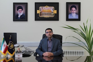 A man sits at an office desk with a reflective surface. There are three framed pictures on the wall behind him, with one picture each of two different men and a central image depicting an architectural structure. To the left on the desk, small flags in stands are visible, along with a telephone and some papers. A large plant is positioned to the right of the desk.