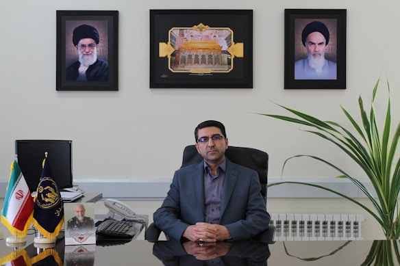 A man sits at an office desk with a reflective surface. There are three framed pictures on the wall behind him, with one picture each of two different men and a central image depicting an architectural structure. To the left on the desk, small flags in stands are visible, along with a telephone and some papers. A large plant is positioned to the right of the desk.
