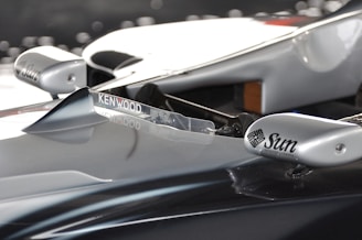 A close-up of a sleek, metallic surface of a racing car, featuring sponsor logos from Kenwood and Sun Microsystems. The surface appears aerodynamic with smooth curves and a polished finish.
