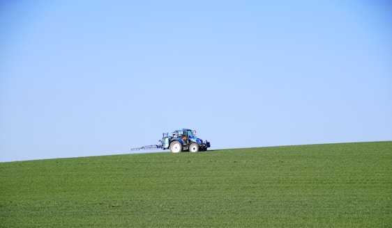A green tractor working in a lush rural field under a clear blue sky.