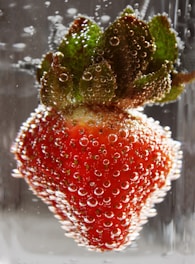 A vibrant red strawberry is fully submerged in carbonated water, with bubbles clinging to its surface, enhancing the texture and color. The green leaves are detailed with numerous small bubbles, creating a refreshing and dynamic visual effect.