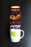 Various dried fruit slices and other ingredients are arranged in a vertical line above a white mug with a green leaf decoration and a blue zig-zag pattern. These ingredients include a slice of dried ginger, red fruit rings, dark dried fruit, brown slices, red petals, and small white cubes.