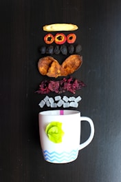 Various dried fruit slices and other ingredients are arranged in a vertical line above a white mug with a green leaf decoration and a blue zig-zag pattern. These ingredients include a slice of dried ginger, red fruit rings, dark dried fruit, brown slices, red petals, and small white cubes.