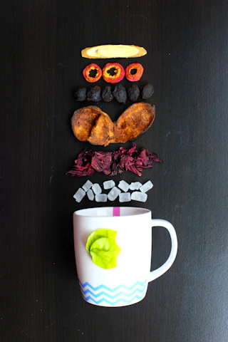 Various dried fruit slices and other ingredients are arranged in a vertical line above a white mug with a green leaf decoration and a blue zig-zag pattern. These ingredients include a slice of dried ginger, red fruit rings, dark dried fruit, brown slices, red petals, and small white cubes.