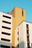 Modern architectural buildings with clean lines and flat surfaces against a vibrant blue sky. The structures feature a combination of white and mustard-colored facades, creating a striking contrast. Large rectangular windows are seen on the buildings, with shadows softly cast across the surfaces.