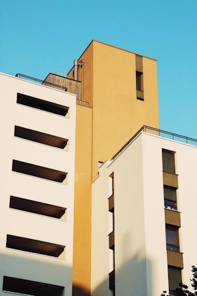 Modern architectural buildings with clean lines and flat surfaces against a vibrant blue sky. The structures feature a combination of white and mustard-colored facades, creating a striking contrast. Large rectangular windows are seen on the buildings, with shadows softly cast across the surfaces.