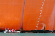 A small boat with people wearing red life jackets sails alongside a massive, orange-painted ship hull. The hull features a series of vertical numbers indicating draft markings. The water below is a bright blue-green, contrasting with the vibrant color of the ship.