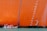 A small boat with people wearing red life jackets sails alongside a massive, orange-painted ship hull. The hull features a series of vertical numbers indicating draft markings. The water below is a bright blue-green, contrasting with the vibrant color of the ship.