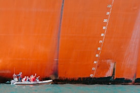 A small boat with people wearing red life jackets sails alongside a massive, orange-painted ship hull. The hull features a series of vertical numbers indicating draft markings. The water below is a bright blue-green, contrasting with the vibrant color of the ship.