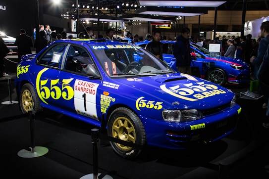 A blue Subaru rally car is displayed at an indoor exhibition. The vehicle features prominent yellow '555' branding and various sponsor logos. Its style is classic and sporty, drawing attention from onlookers in the background.