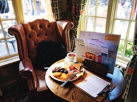 A cozy setting with a leather armchair next to a round wooden table. On the table, there is a full English breakfast with items like sausages, beans, and toast, accompanied by a cup of tea. Various menus and flyers are propped up on the table. Sunlight streams through nearby windows, creating a warm and inviting atmosphere.