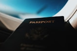 A close-up of a passport leaning against the window of an airplane, with an aerial view of the sky and clouds in the background