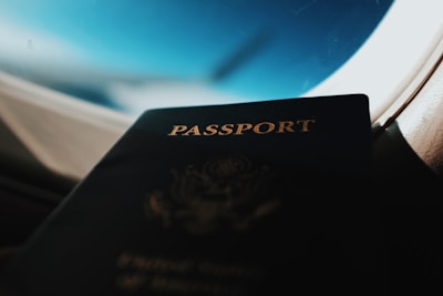 A close-up of a passport leaning against the window of an airplane, with an aerial view of the sky and clouds in the background