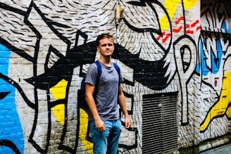 A young person stands in front of a wall covered in vibrant, abstract graffiti art. The mural includes bold black lines and patches of primary colors like yellow, red, and blue against a white background. The person is casually dressed in a gray t-shirt and blue jeans, carrying a backpack and looking confidently at the camera.