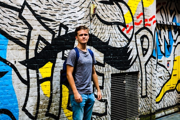 A young person stands in front of a wall covered in vibrant, abstract graffiti art. The mural includes bold black lines and patches of primary colors like yellow, red, and blue against a white background. The person is casually dressed in a gray t-shirt and blue jeans, carrying a backpack and looking confidently at the camera.