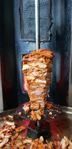 Close-up photo of a sizzling, freshly grilled chicken döner rotating on a vertical spit with flames in the background.