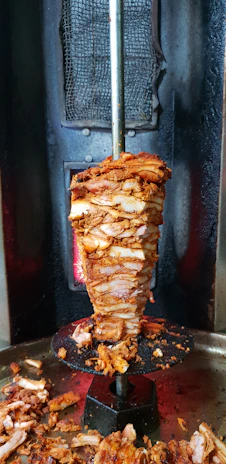 Close-up of a juicy, freshly sliced chicken döner rotating on a vertical spit with flames in the background.