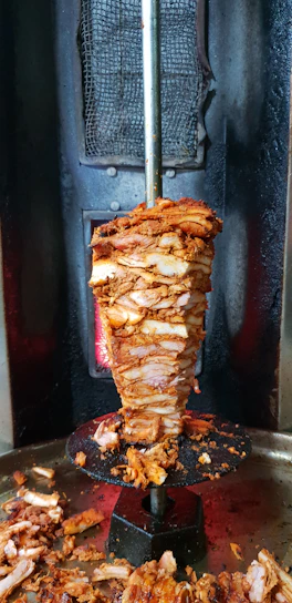 Close-up photo of a sizzling, freshly grilled chicken döner rotating on a vertical spit with flames in the background.