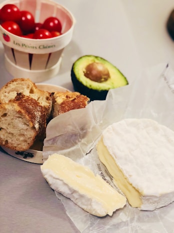 A composition of fresh food items including brie cheese on wax paper, a sliced baguette, a halved avocado displaying its seed, and a bowl filled with cherry tomatoes.