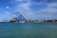 Large blue cranes stand prominently against a partly cloudy sky at a bustling port, with numerous stacked shipping containers visible along the dock. The calm sea in the foreground is dotted with small boats, creating a scene of industrial activity and maritime transport.