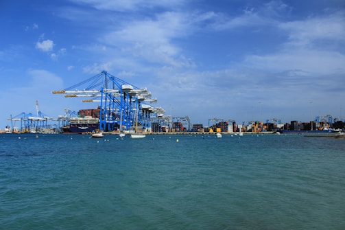 Large blue cranes stand prominently against a partly cloudy sky at a bustling port, with numerous stacked shipping containers visible along the dock. The calm sea in the foreground is dotted with small boats, creating a scene of industrial activity and maritime transport.