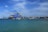 Large blue cranes stand prominently against a partly cloudy sky at a bustling port, with numerous stacked shipping containers visible along the dock. The calm sea in the foreground is dotted with small boats, creating a scene of industrial activity and maritime transport.