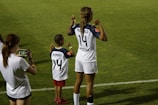 A soccer player and a child stand on a grassy field showing the backs of their jerseys with the numbers 14 and 24. The child wears red shorts and stands with fists raised, mimicking the stance of the soccer player. Another person stands nearby, capturing the moment on a smartphone.