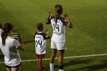 A soccer player and a child stand on a grassy field showing the backs of their jerseys with the numbers 14 and 24. The child wears red shorts and stands with fists raised, mimicking the stance of the soccer player. Another person stands nearby, capturing the moment on a smartphone.