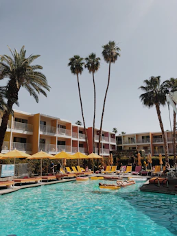 A vibrant outdoor pool area is surrounded by colorful apartment buildings under a clear sky. Tall palm trees rise above the scene, and numerous yellow lounge chairs and umbrellas are arranged around the pool. People are relaxing and enjoying the sunny day, some floating on inflatable rafts in the pool.