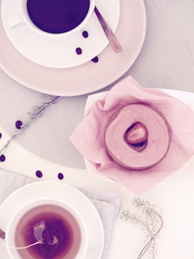 A light-colored table setup featuring a cup of coffee on a saucer, surrounded by coffee beans and a delicate branch. Nearby, there is a cup of tea with a tea bag inside on a cloth napkin. A pink frosted dessert with a strawberry slice on top is placed on pink tissue paper.