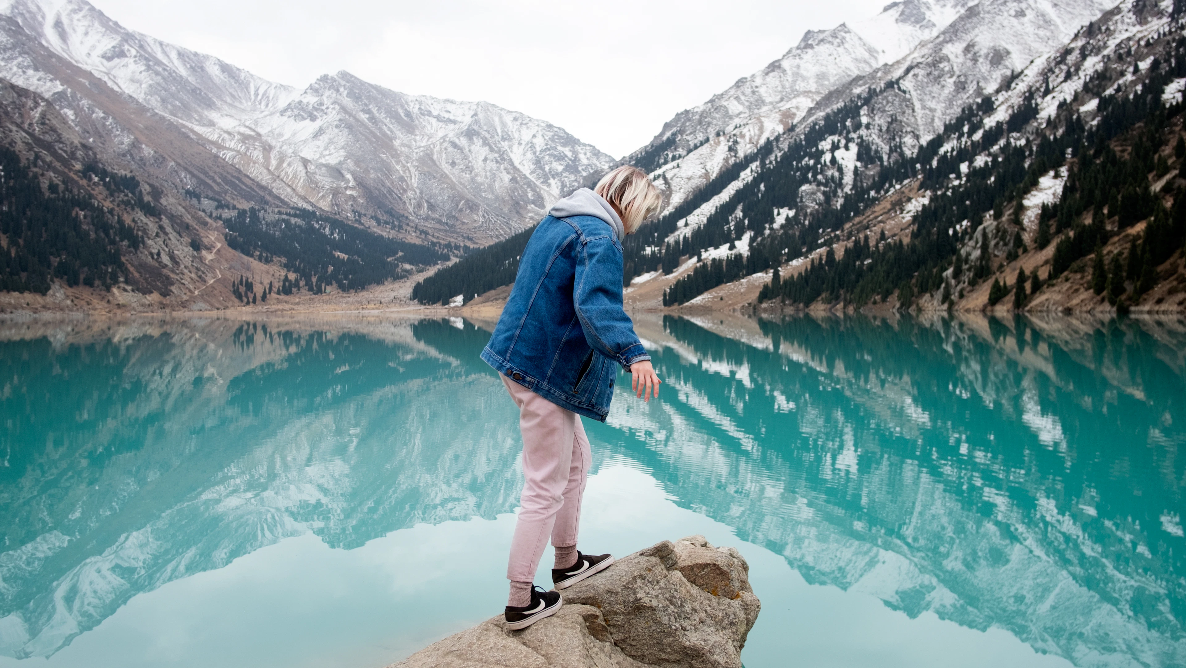 A traveler in a denim jacket stands on a boulder above Big Almaty Lake's mirror-still turquoise water, ringed by snowy peaks.