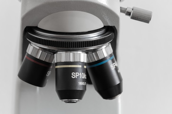 A close-up view of a microscope lens system with multiple objective lenses set on a rotating turret. The lenses are labeled with specifications and are mounted on a silver and black frame. The background is out of focus, highlighting the detailing on the microscope.