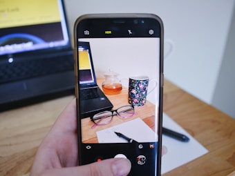 A hand holds a smartphone with its camera app open, capturing an image of a desk setup. The desk has a laptop, a pot of tea, a floral mug, and a pair of glasses resting on a piece of paper with a pen. The background is minimal, with focus on the objects displayed.