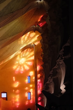 Intricate architectural features with dynamic lighting provide a colorful display. The structure is illuminated with warm tones of yellow, orange, and red light, creating vibrant reflections on the walls. Several small windows interrupt the play of light, integrated into the complex, sculptural details of the design.